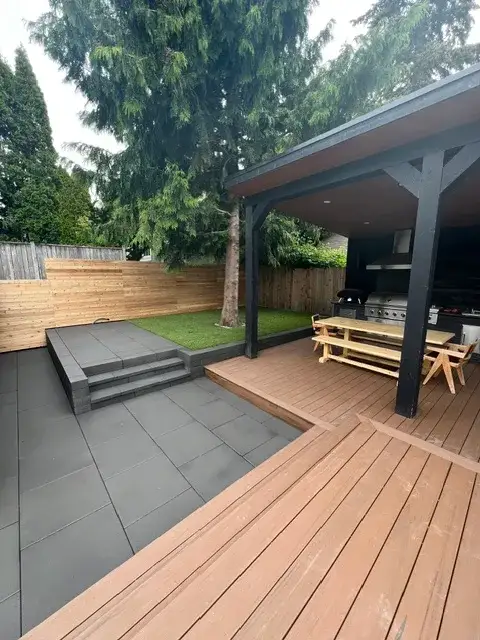 Covered outdoor kitchen with a built-in grill and picnic tables beside a warm-toned deck, grey slab patio and artificial turf lawn in a Magnolia back garden