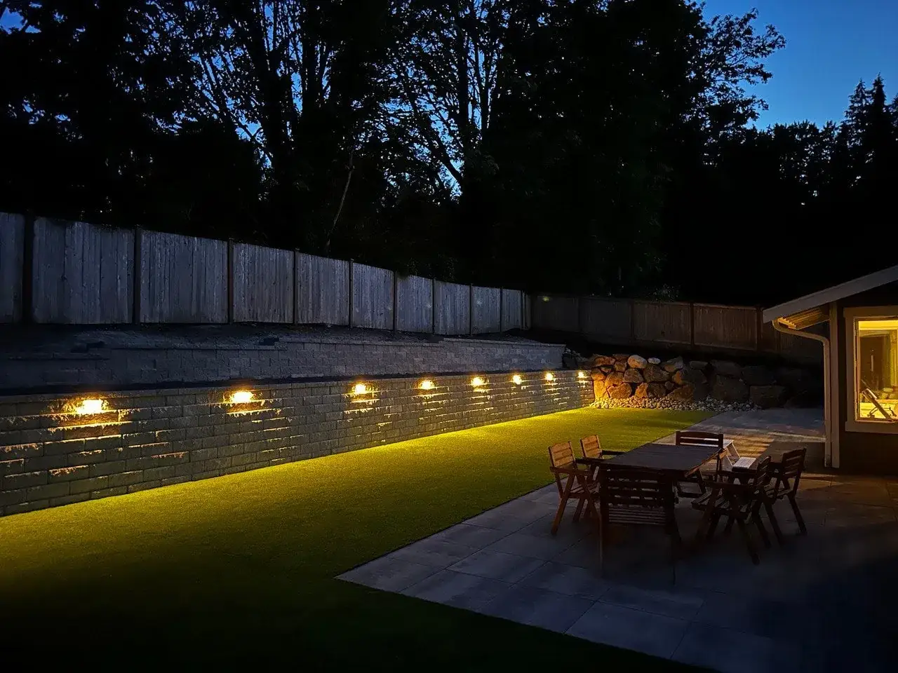 Landscape lighting washing a retaining wall above a lit patio at dusk
